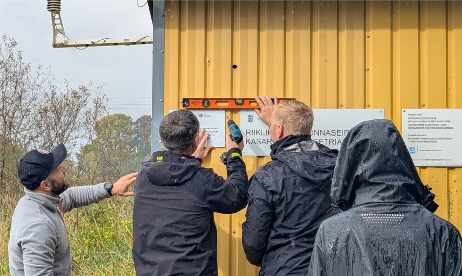 Neli inimest Kasari hüdromeetriajaama hoone seinale WMO tunnustuse plaati asetamas. 2 neist paigaldavad plaati, ühe käes on puur, teine hoiab paigal loodi. Kaks vaatavad pealt.